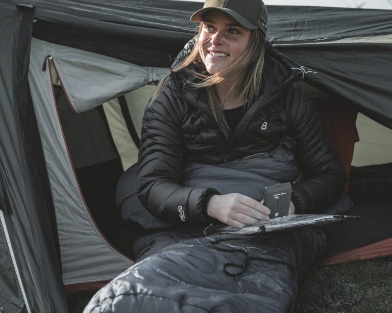 Easy Camp Falcon II Mummy -1°C 2 Uma mulher senta-se num Easy Camp Falcon II Mummy -1°C Saco de Dormir em frente a uma tenda.