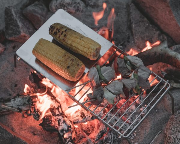 O Campfire Grill da marca easy camp dobrável, à luz de uma fogueira com alimentos grelhados O Campfire Grill da marca easy camp dobrável, à luz de uma fogueira com alimentos grelhados