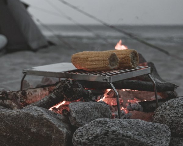 O Campfire Grill da marca easy camp dobrável, à luz de uma fogueira com alimentos grelhados O Campfire Grill da marca easy camp dobrável, à luz de uma fogueira com alimentos grelhados