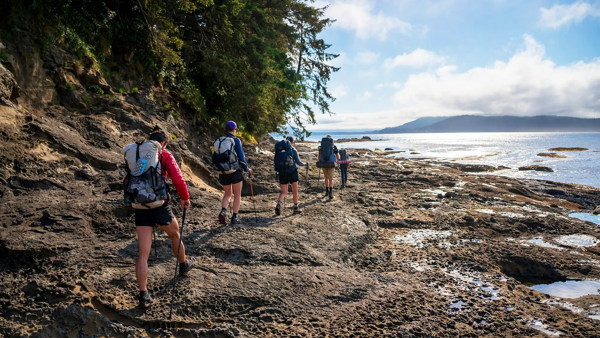 Pessoa a caminhar num trilho de montanha durante o verão com mochila de caminhada, rodeada por paisagens naturais e céu limpo.